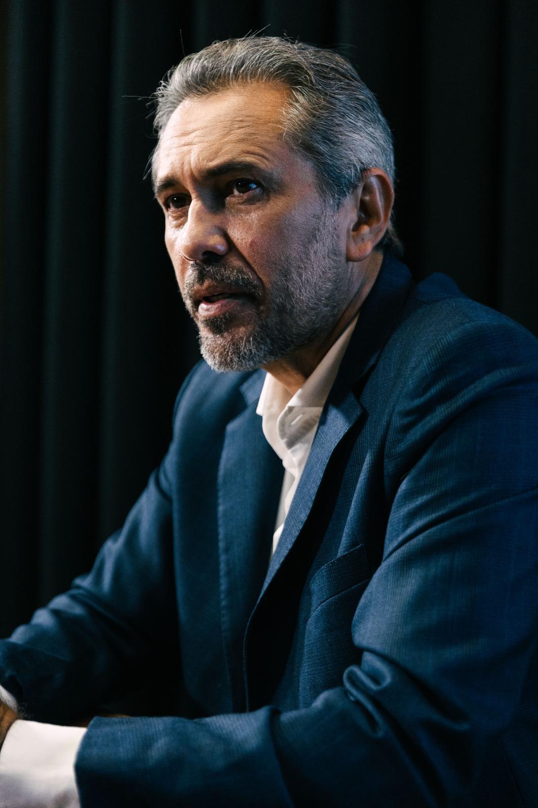 Elmano de Freitas de terno azul, sentado à mesa durante entrevista. Ele mantém as mãos apoiadas sobre a mesa, com um copo de água e uma caneca ao lado, diante de um fundo escuro com cortinas.”