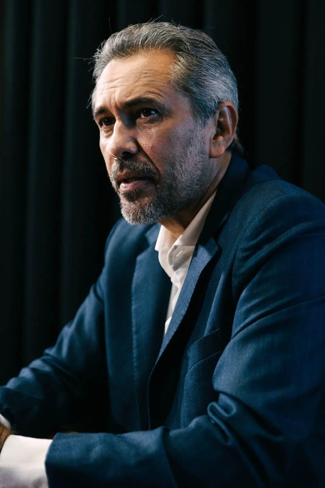 Elmano de Freitas de terno azul, sentado à mesa durante entrevista. Ele mantém as mãos apoiadas sobre a mesa, com um copo de água e uma caneca ao lado, diante de um fundo escuro com cortinas.”