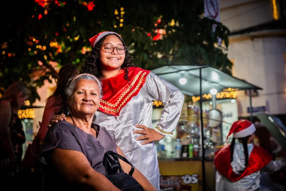 Na imagem, uma foto noturna em ambiente externo. Em primeiro plano e sentada, está Dona Marilene, uma mulher idosa de pele clara e cabelo grisalho, sorrindo gentilmente para a câmera. Ela usa uma blusa escura de manga curta. Atrás dela, em pé, está Ana Emilly, uma jovem de pele morena, cabelo cacheado e óculos, com um sorriso aberto e a mão na cintura. Ela veste um traje de Natal composto por uma bata branca e um xale vermelho e dourado, além de um gorro de Papai Noel vermelho e branco. Elas estão abraçadas, com o braço de Dona Marilene no ombro de Ana Emilly. O fundo está desfocado (bokeh), com luzes de Natal amarelas e vermelhas e a silhueta de outras pessoas e uma barraca iluminada.