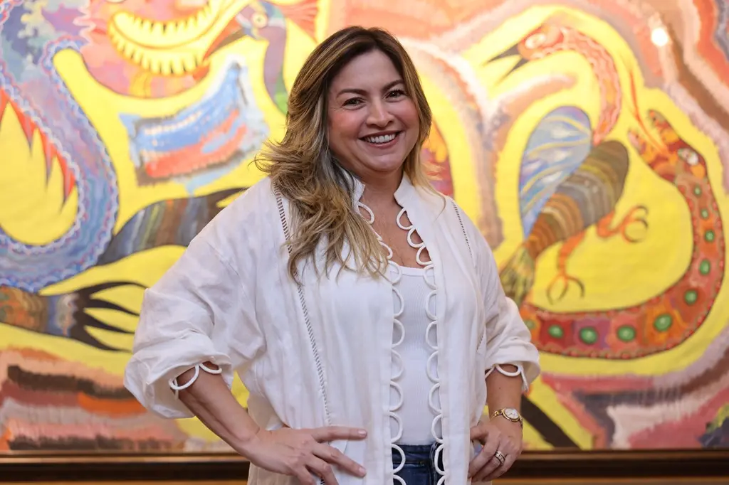 Ana Quezado, mulher loira, com roupa branca, sorri para a foto. Ela aparenta estar em uma exposição.