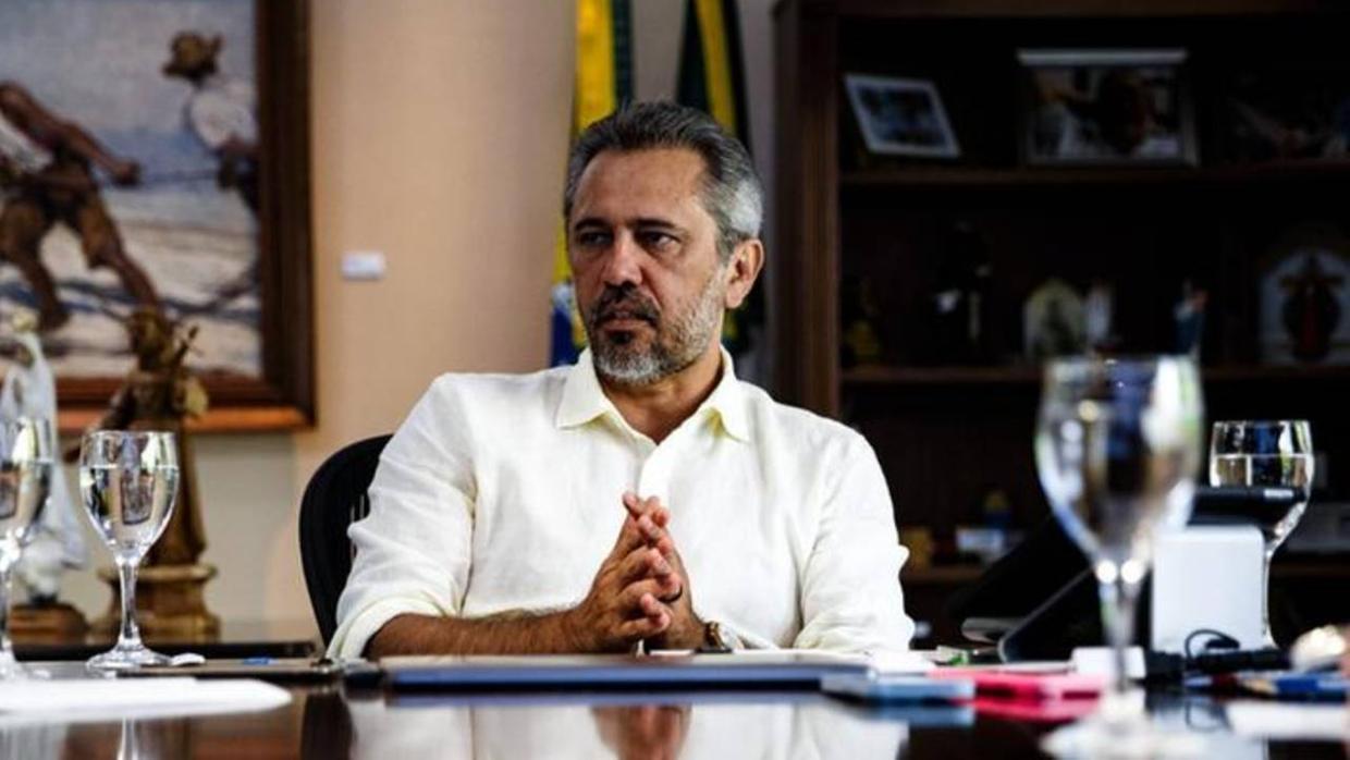 Elmano de Freitas de camisa clara, sentado à mesa em um gabinete, com as mãos entrelaçadas à frente. Sobre a mesa há documentos, um telefone e taças de água; ao fundo, estantes e obras decorativas em ambiente institucional.