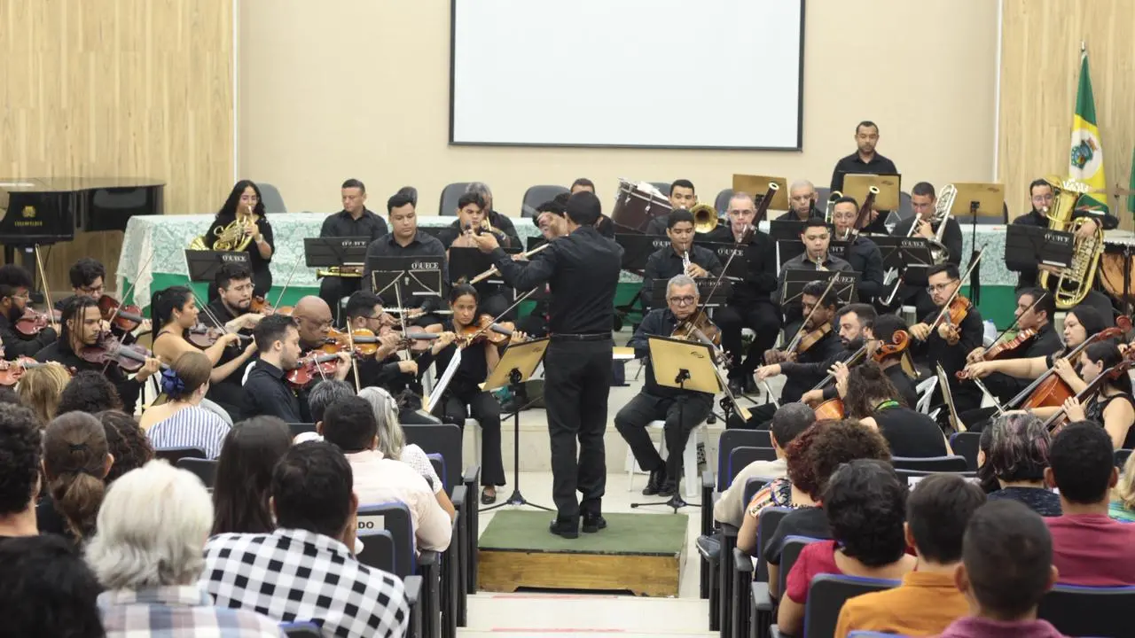Vista da plateia de uma orquestra sinfônica apresentando-se em um auditório. No centro, um maestro de costas, vestido de preto, rege o grupo sobre um pequeno tablado. Os músicos, também vestidos de preto, estão organizados em semicírculo. O ambiente possui paredes de madeira clara e um telão branco vazio ao centro. Em primeiro plano, as cabeças dos espectadores sentados em poltronas azuis aparecem de costas, desfocadas.