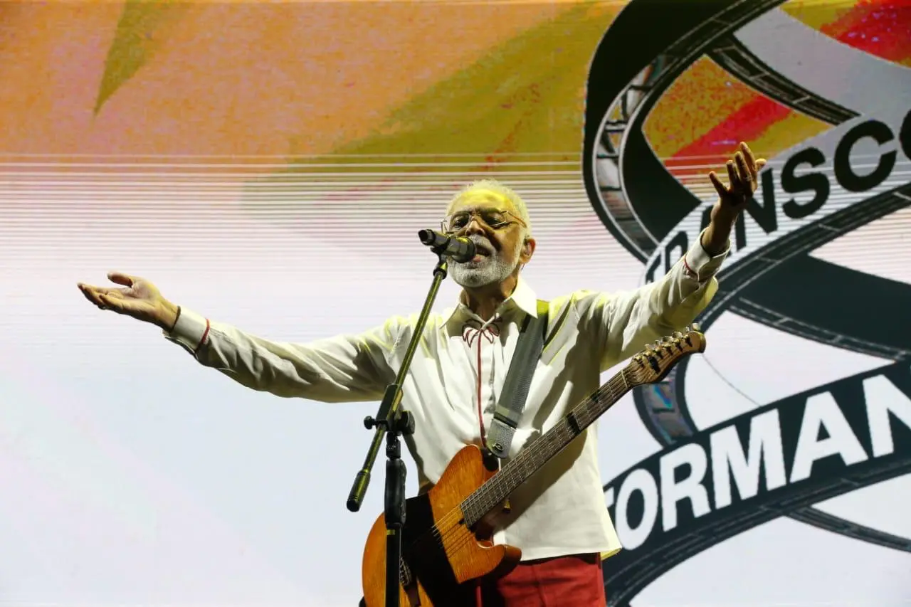 Cantor Gilberto Gil se apresenta no palco com violão, braços abertos diante do público, durante show com telão ao fundo.