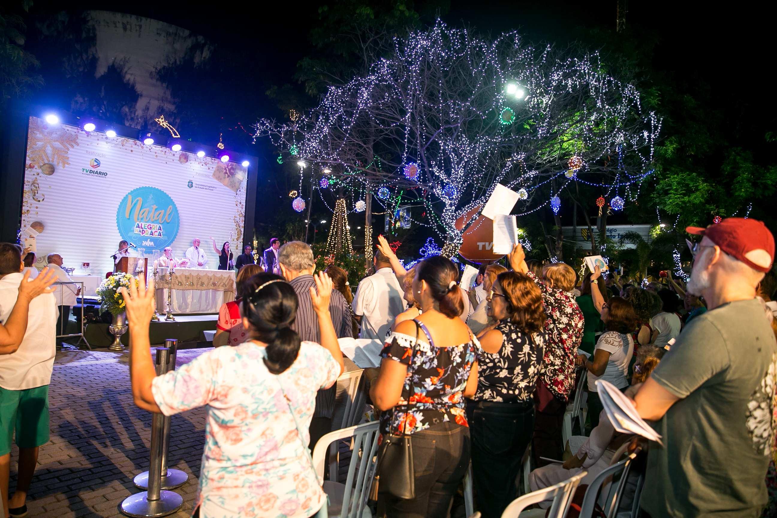 A imagem noturna capta uma Missa de Natal ao ar livre na Praça da Imprensa, mostrando uma multidão reunida em frente a um palco iluminado. O evento destaca-se pela sua atmosfera festiva e religiosa, com o palco exibindo um telão com o tema 
