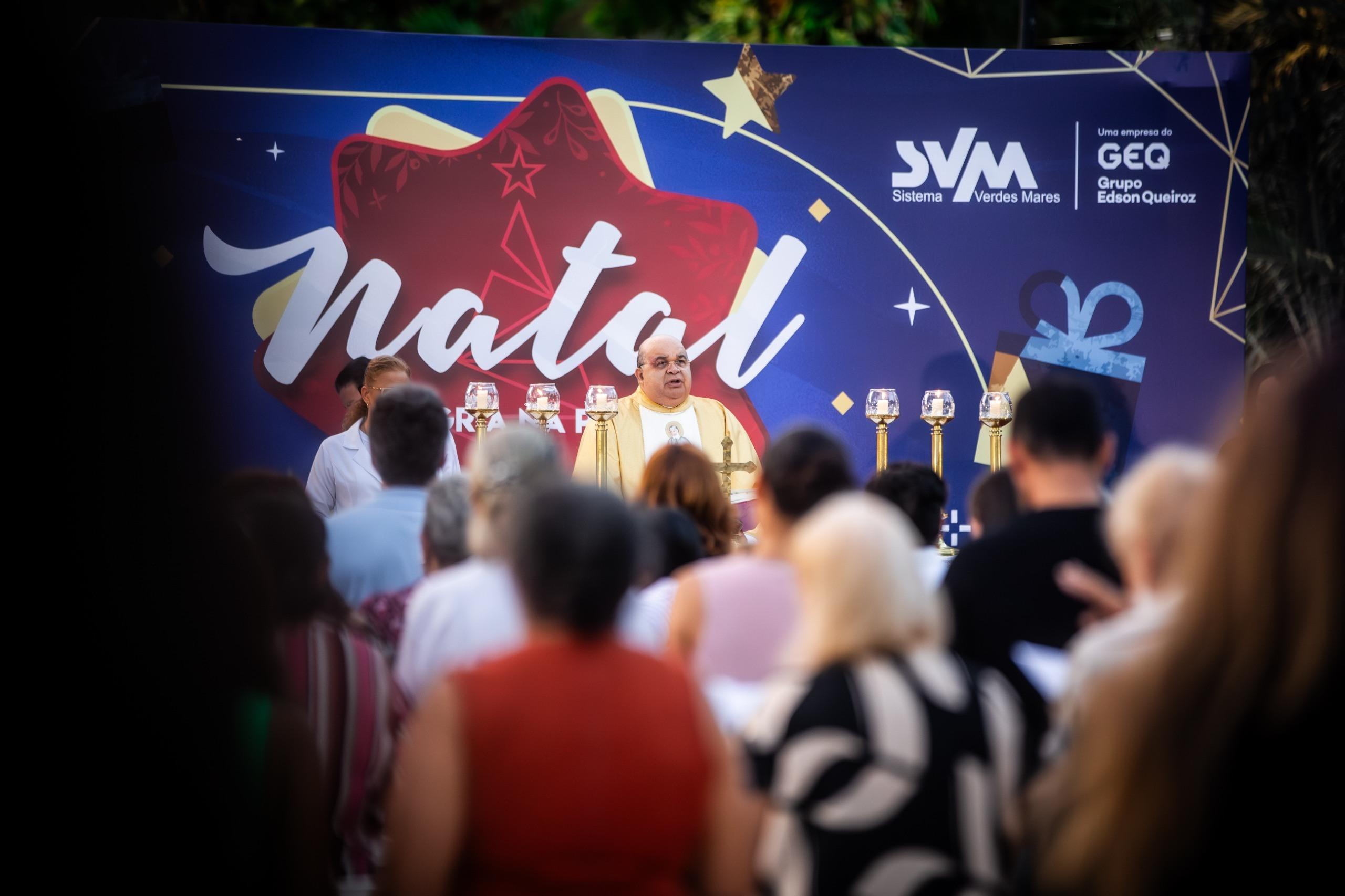 A imagem noturna capta uma Missa de Natal ao ar livre na Praça da Imprensa, mostrando uma multidão reunida em frente a um palco iluminado. O evento destaca-se pela sua atmosfera festiva e religiosa, com o palco exibindo um telão com o tema 