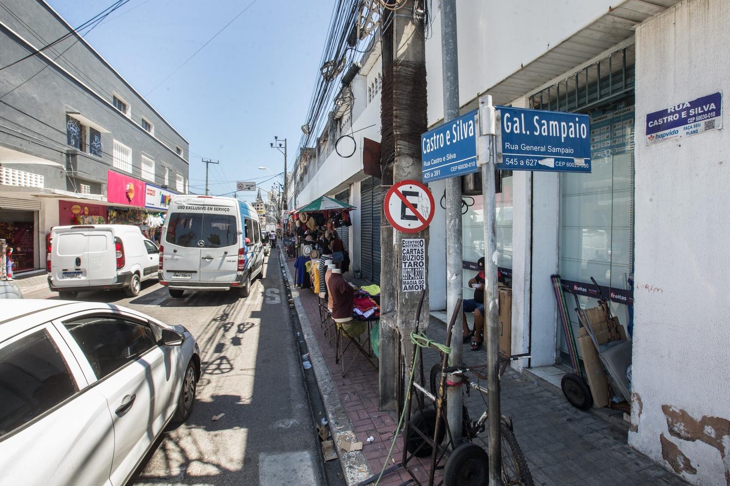 Foto que contém placa do cruzamento das ruas Castro e Silva e General Sampaio, no Centro de Fortaleza. Ao fundo, comércio formal e informal na via.
