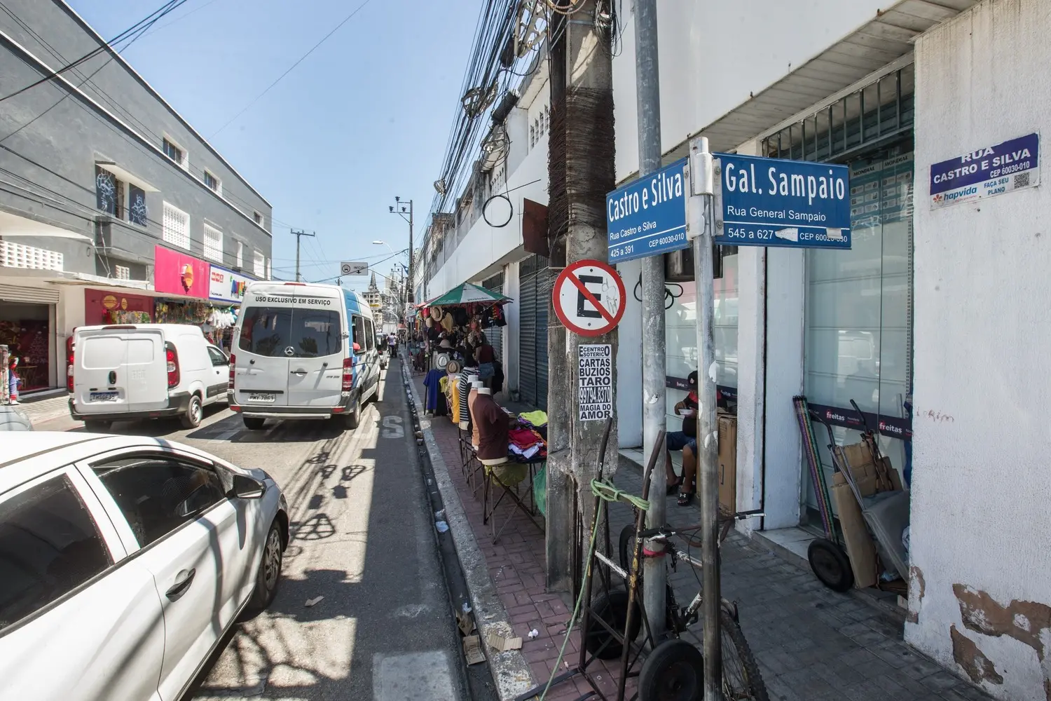 Foto que contém placa do cruzamento das ruas Castro e Silva e General Sampaio, no Centro de Fortaleza. Ao fundo, comércio formal e informal na via.
