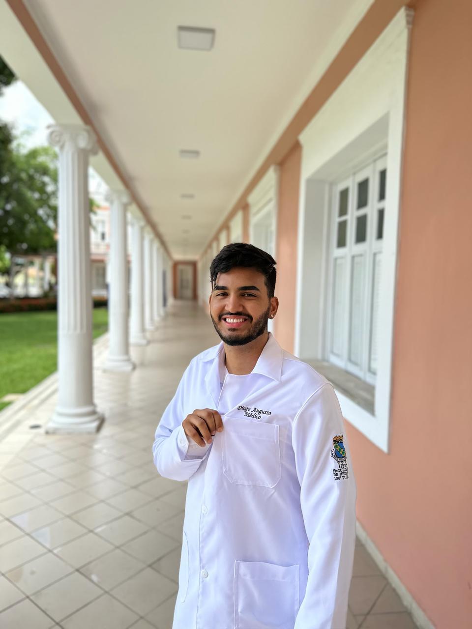 Imagem mostra Diogo Augusto, primeiro médico quilombola formado pela UFC, no corredor da universidade, vestindo jaleco e sorrindo.