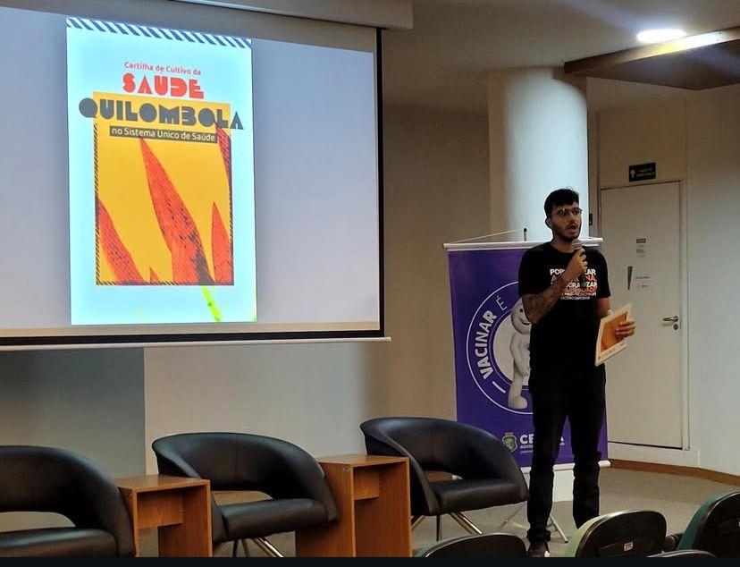Imagem mostra estudante Diogo todo vestido de preto em um palco durante palestra sobre Saúde Quilombola na UFC.