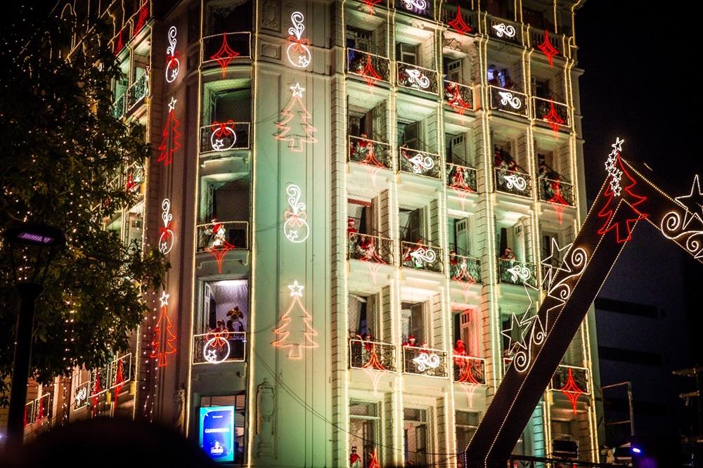 Na imagem, foto noturna de um edifício antigo e clássico, iluminado com decorações de Natal brilhantes. As luzes de LED brancas e vermelhas formam desenhos de árvores de Natal, estrelas e sinos ao redor das janelas e varandas. O edifício tem vários andares e sacadas com pessoas observando as decorações. Uma grande estrutura decorativa em forma de arco ou ponte, também coberta por luzes, se estende para a direita, e uma árvore com luzes cintilantes fica à esquerda. A cena está escura ao redor, destacando a iluminação festiva do prédio.