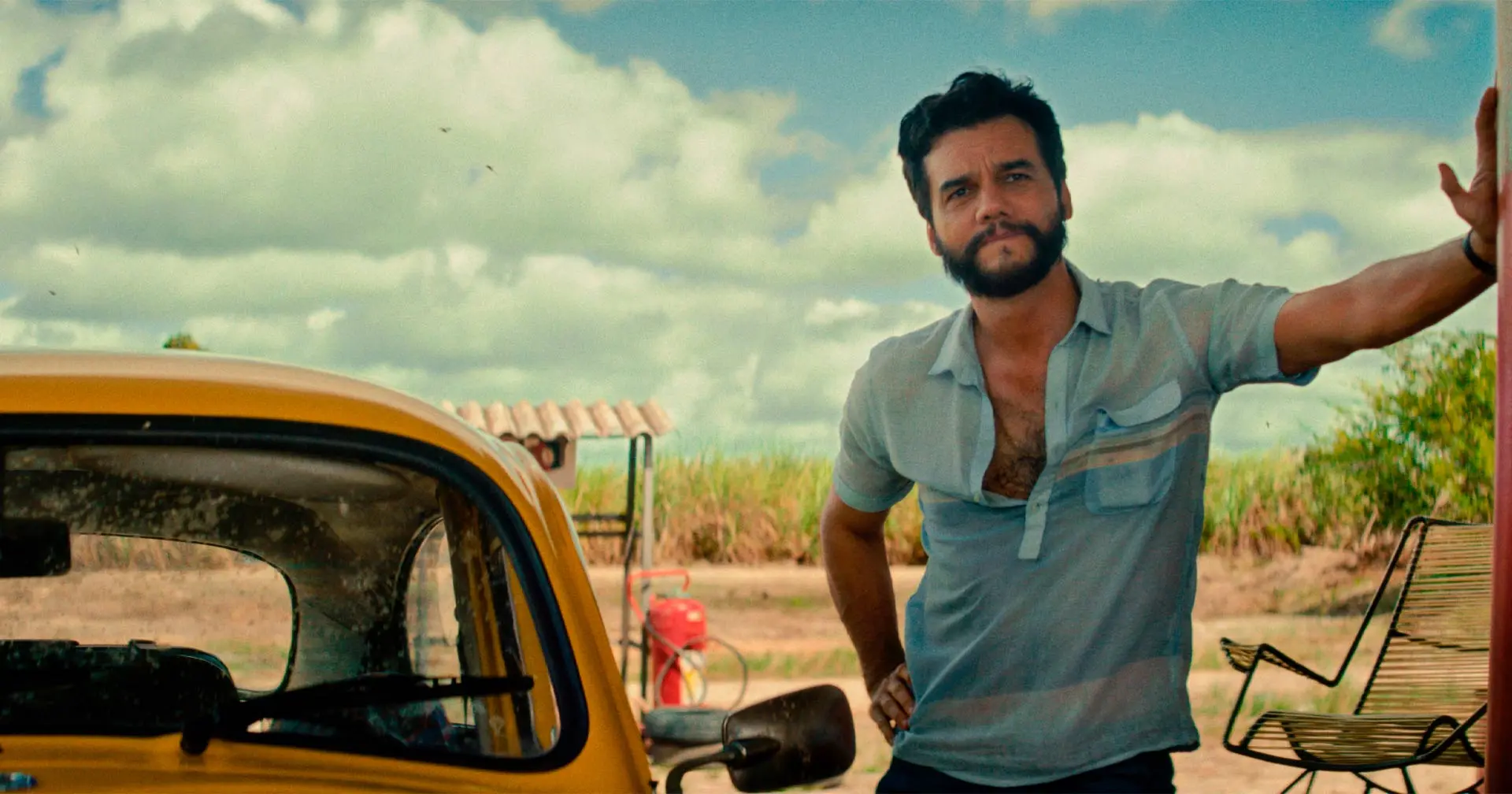 Homem encostado em carro amarelo antigo em cenário rural com céu azul e canavial ao fundo. Cena do filme O Agente Secreto, com Wagner Moura.