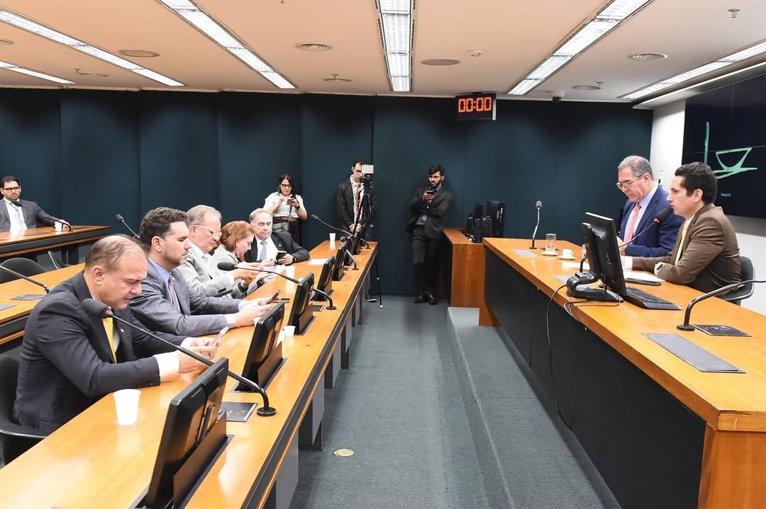 Sala de audiência ou reunião institucional com mesas de madeira dispostas paralelamente. À direita, dois homens de terno sentados à mesa principal falam ao microfone e consultam documentos em frente a monitores. À esquerda, vários homens e mulheres também de terno estão sentados em fileiras, alguns olhando para celulares ou telas. Ao fundo, há jornalistas e cinegrafistas registrando o evento, um relógio digital marcando “00:00” e um painel com símbolo da Justiça. O ambiente é formal, com iluminação fluorescente e clima de sessão oficial.
