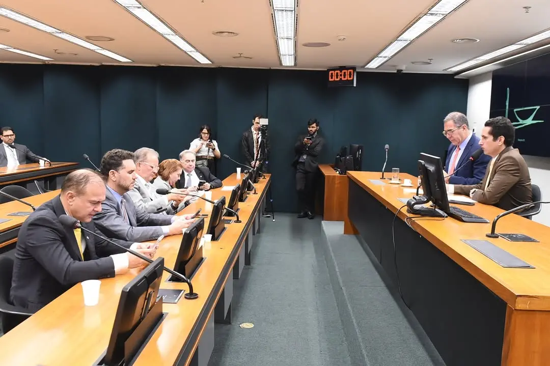 Sala de audiência ou reunião institucional com mesas de madeira dispostas paralelamente. À direita, dois homens de terno sentados à mesa principal falam ao microfone e consultam documentos em frente a monitores. À esquerda, vários homens e mulheres também de terno estão sentados em fileiras, alguns olhando para celulares ou telas. Ao fundo, há jornalistas e cinegrafistas registrando o evento, um relógio digital marcando “00:00” e um painel com símbolo da Justiça. O ambiente é formal, com iluminação fluorescente e clima de sessão oficial.
