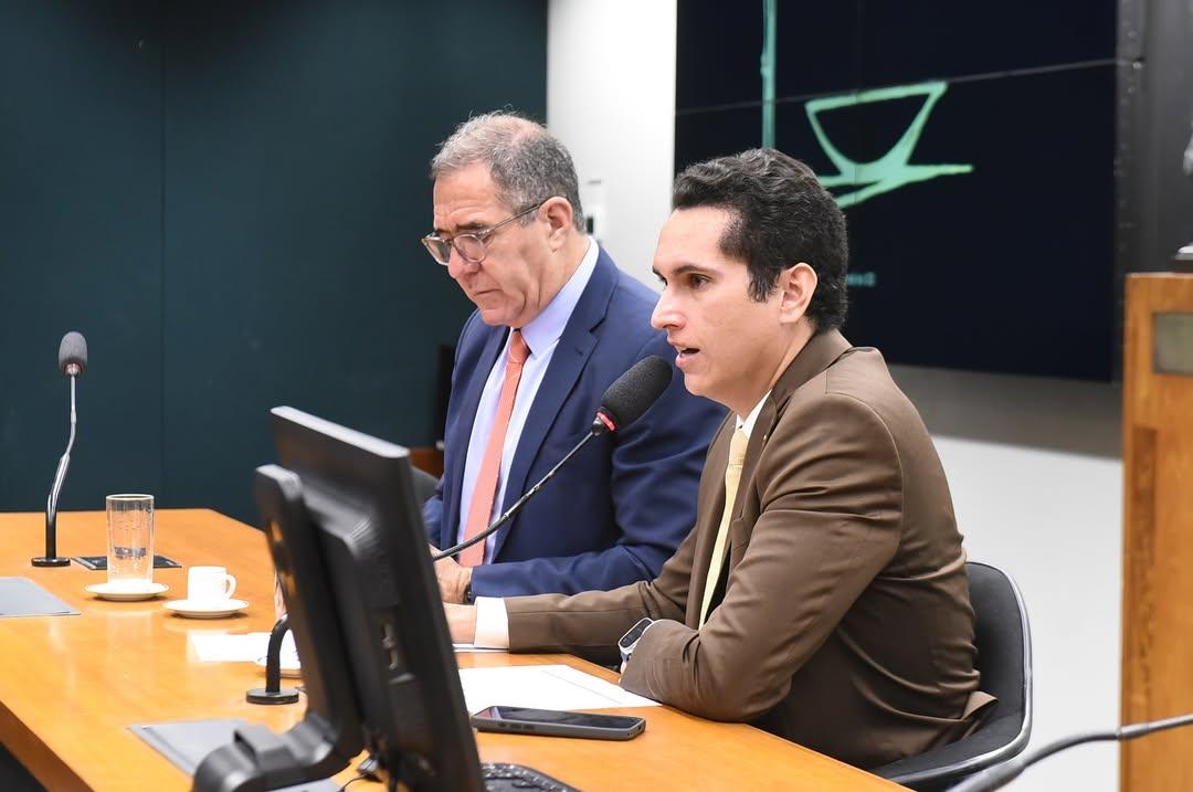Dois homens de terno sentados à mesa principal de uma sala de audiência. O homem à frente, com terno marrom, fala ao microfone enquanto olha para a frente, com expressão concentrada. Ao lado dele, um homem de terno azul e gravata clara observa documentos em frente a um monitor. Sobre a mesa há monitores, microfones, um copo de água, uma xícara de café e um celular. Ao fundo, aparece um painel com símbolo da Justiça, reforçando o ambiente formal e institucional.
