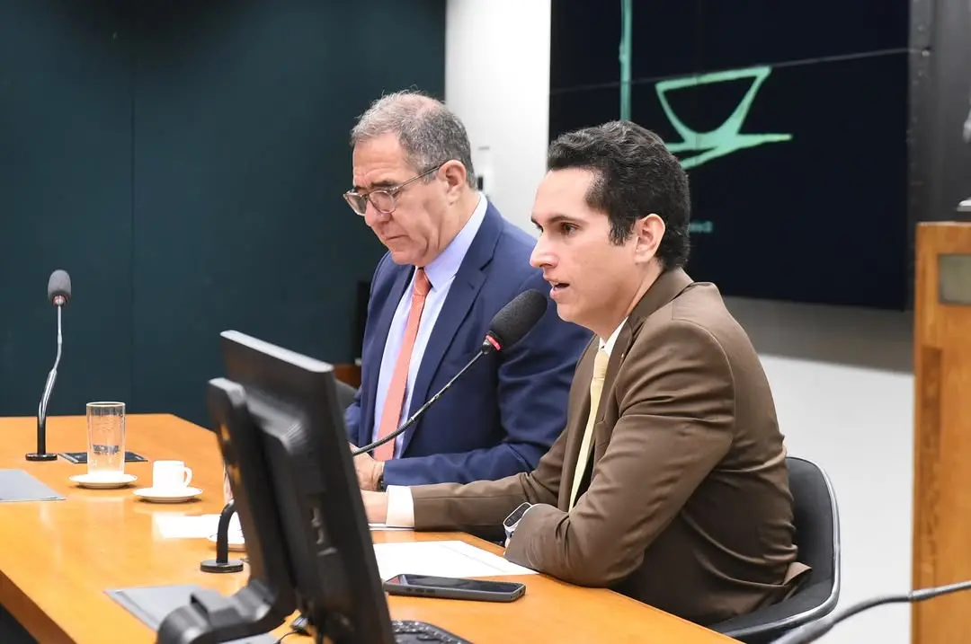 Dois homens de terno sentados à mesa principal de uma sala de audiência. O homem à frente, com terno marrom, fala ao microfone enquanto olha para a frente, com expressão concentrada. Ao lado dele, um homem de terno azul e gravata clara observa documentos em frente a um monitor. Sobre a mesa há monitores, microfones, um copo de água, uma xícara de café e um celular. Ao fundo, aparece um painel com símbolo da Justiça, reforçando o ambiente formal e institucional.
