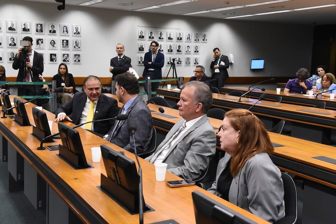 Sala de comissão legislativa com mesas de madeira e cadeiras pretas. Em primeiro plano, quatro pessoas de terno estão sentadas lado a lado; duas conversam entre si, enquanto as outras observam a movimentação. Sobre as mesas há microfones, monitores e copos descartáveis. Ao fundo, jornalistas e assessores em pé acompanham a sessão, alguns com câmeras e celulares. Na parede, há quadros com retratos em preto e branco de ex-presidentes da instituição, reforçando o ambiente formal e institucional.
