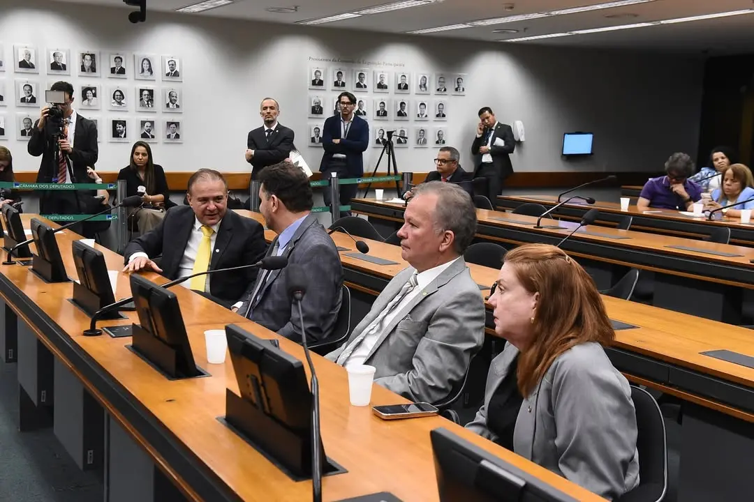 Sala de comissão legislativa com mesas de madeira e cadeiras pretas. Em primeiro plano, quatro pessoas de terno estão sentadas lado a lado; duas conversam entre si, enquanto as outras observam a movimentação. Sobre as mesas há microfones, monitores e copos descartáveis. Ao fundo, jornalistas e assessores em pé acompanham a sessão, alguns com câmeras e celulares. Na parede, há quadros com retratos em preto e branco de ex-presidentes da instituição, reforçando o ambiente formal e institucional.
