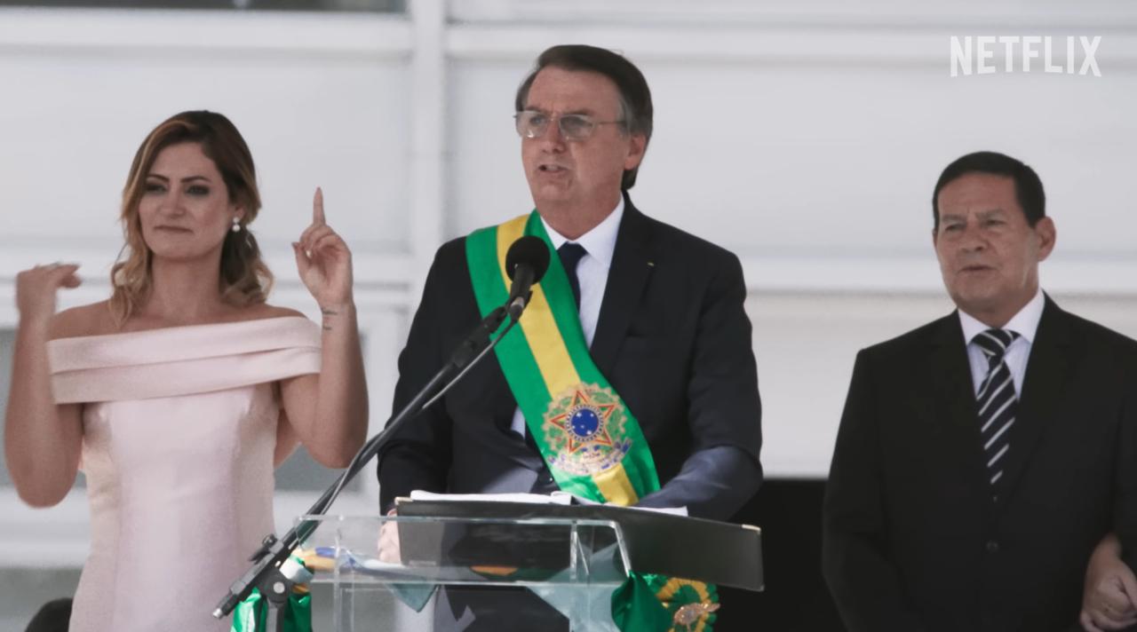 Cerimônia oficial com três pessoas ao lado de um púlpito, incluindo uma figura usando faixa nas cores verde, amarela e azul, em ambiente institucional.