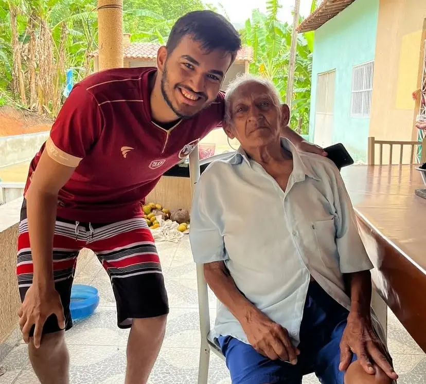 Imagem mostra jovem de 24 anos, Diogo, vestido de camisa e bermuda vermelhas, agachado próximo ao avô de 100 anos, que está sentado vestindo uma camisa social azul clara.