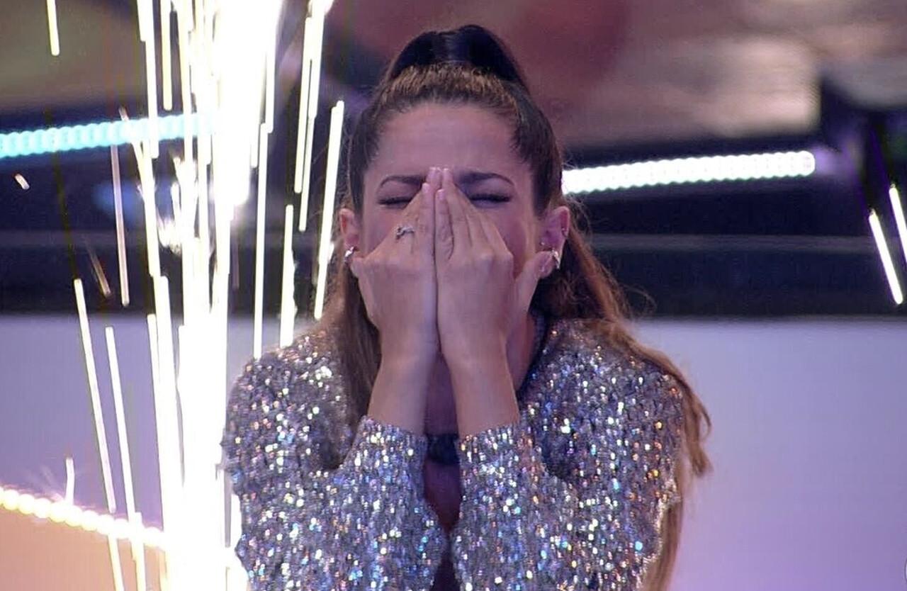 Juliette, mulher branca de cabelos longos, castanhos e lisos presos em um rabo de cavalo, com mãos no rosto e olhos fechados. Ela usa uma roupa prateada brilhante de manga longa e há luzes de celebração ao fundo.