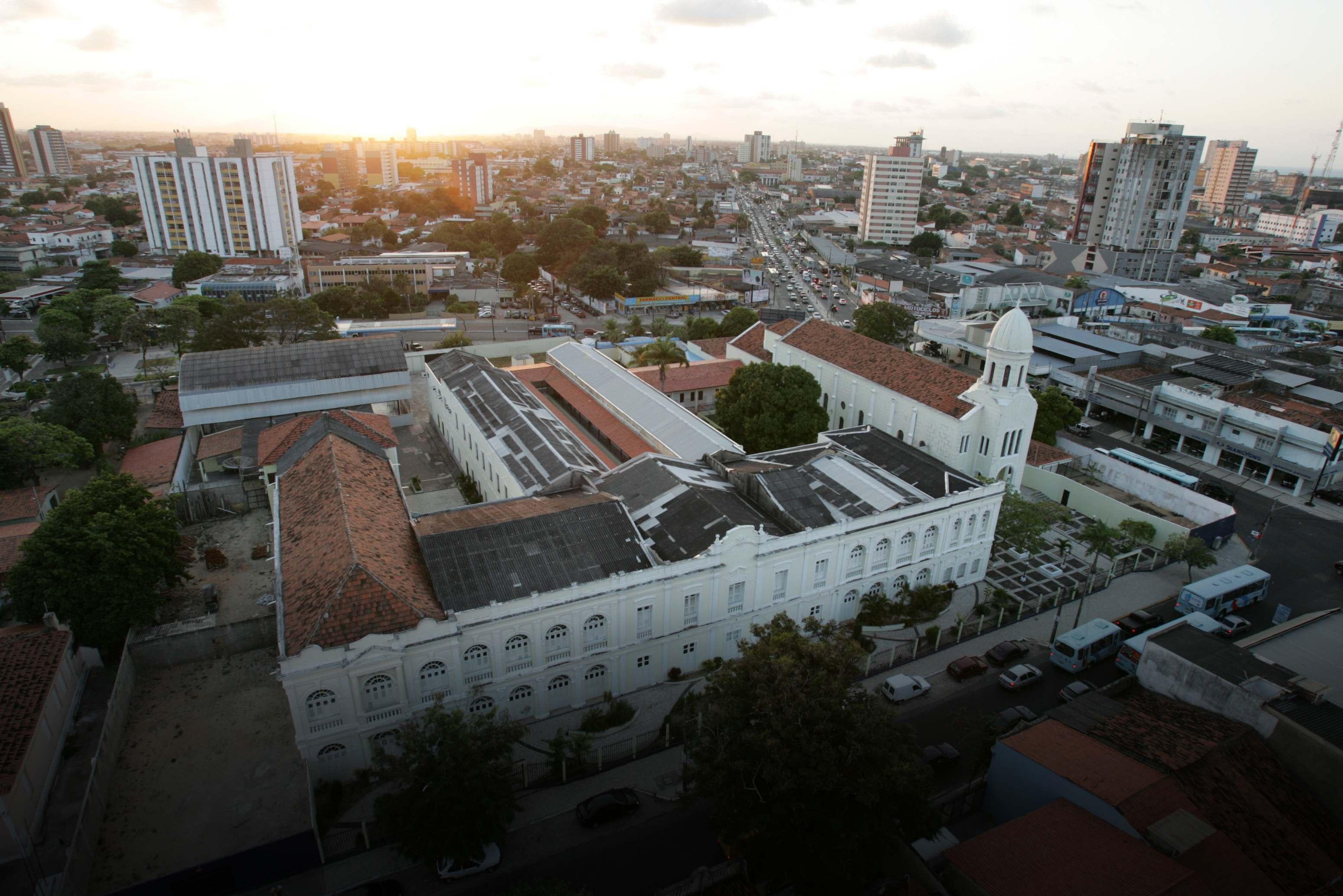 Vista aérea do Colégio das Doroteias em Fortaleza, com sua arquitetura neoclássica branca em destaque no primeiro plano e a densa malha urbana da cidade ao fundo durante o pôr do sol.