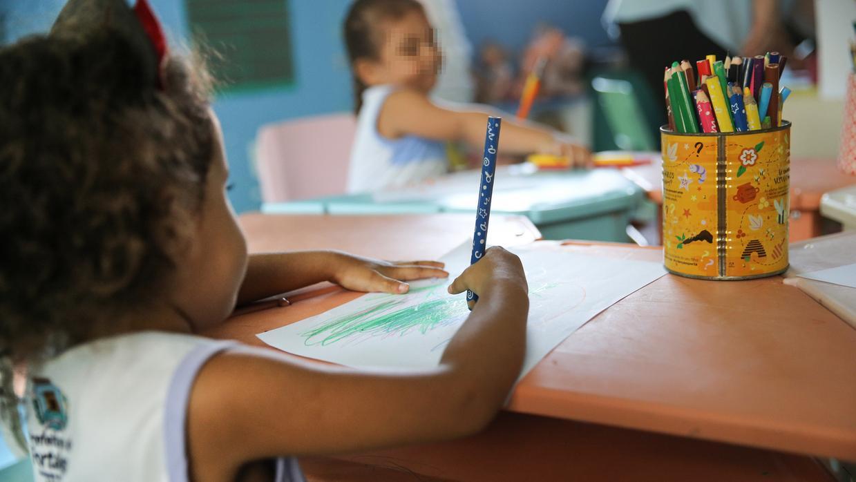 Criança desenhando em creche. À frente, uma mesa laranja e uma lata com lápis de cores.