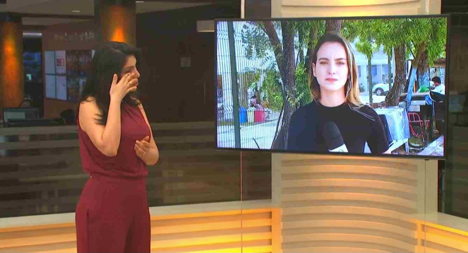 Apresentadora de telejornal em estúdio limpa as lágrimas enquanto observa, em um monitor, uma repórter que também aparece emocionada durante uma transmissão ao vivo.