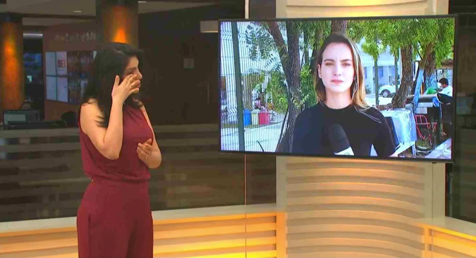 Apresentadora de telejornal em estúdio limpa as lágrimas enquanto observa, em um monitor, uma repórter que também aparece emocionada durante uma transmissão ao vivo.