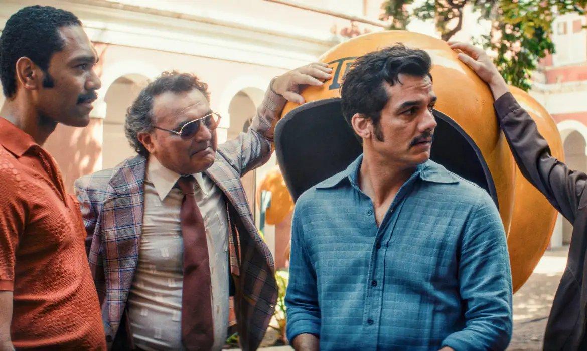 Três homens com expressões sérias, incluindo um com bigode e camisa azul, outro de óculos escuros e terno xadrez, e um de camisa polo laranja, próximos a um orelhão (telefone público) amarelo. A cena remete a um drama histórico ou série.