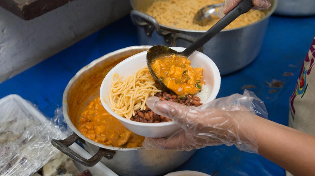 Mãos enluvadas servindo um prato com arroz, feijão, macarrão e frango cozido em uma marmita descartável.