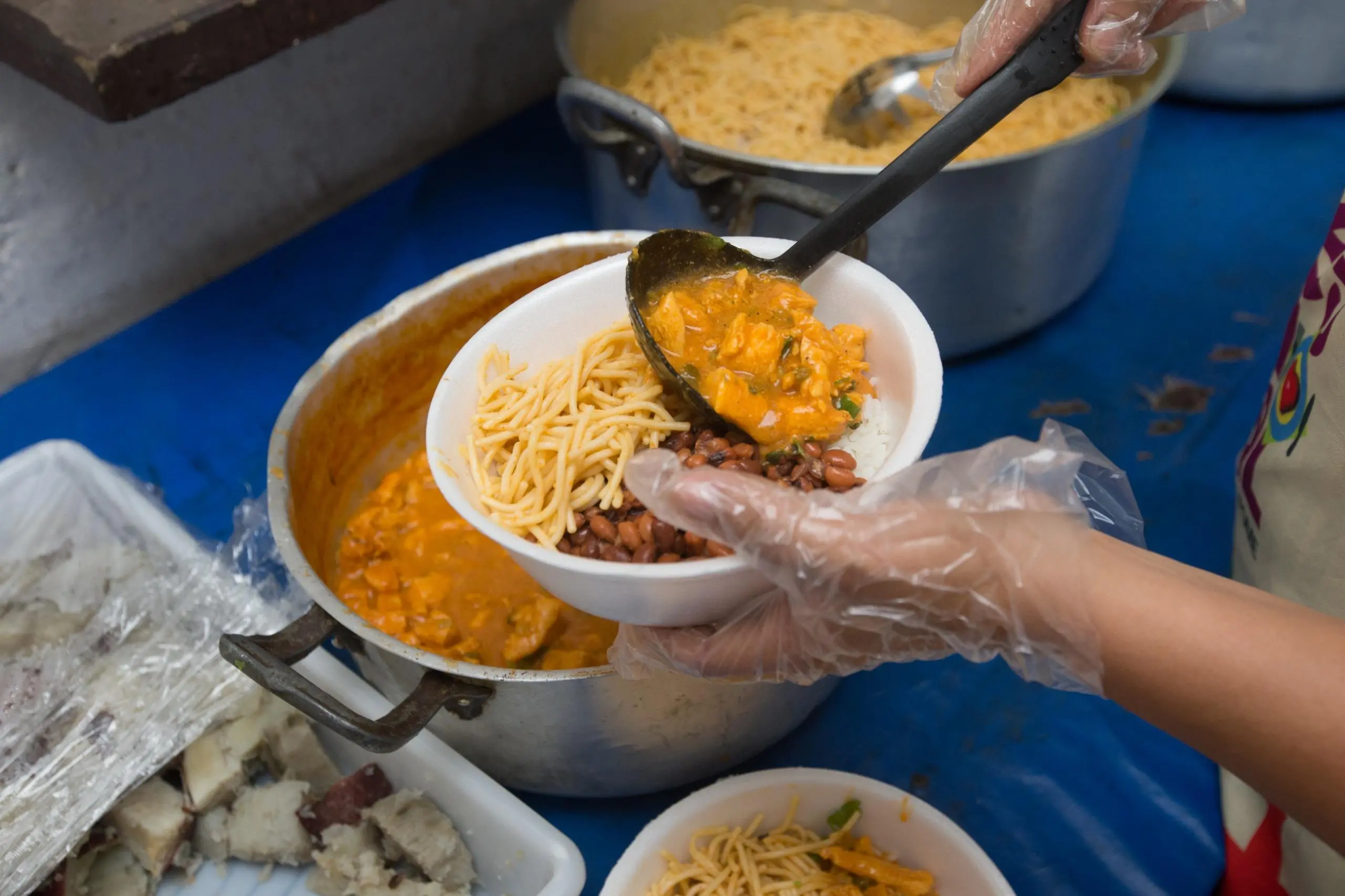 Mãos enluvadas servindo um prato com arroz, feijão, macarrão e frango cozido em uma marmita descartável.