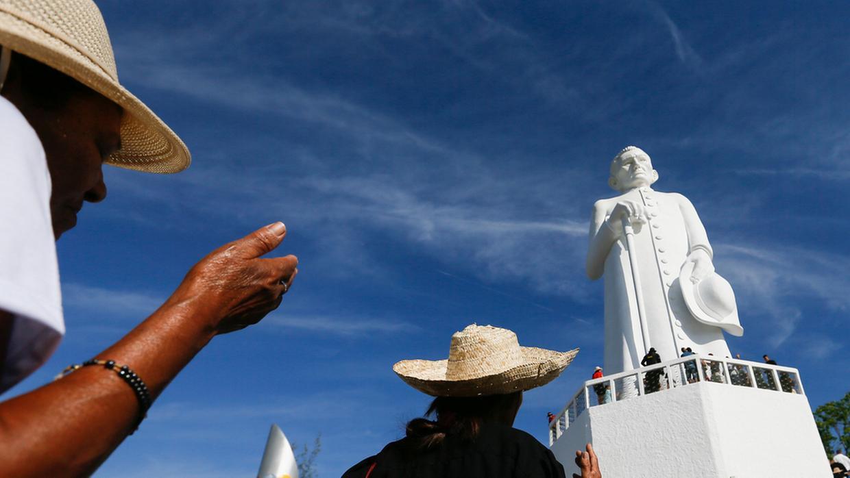 Fiéis usam chapéus de palha e fazem gestos de devoção diante da estátua de Padre Cícero, em Juazeiro do Norte, sob céu azul.