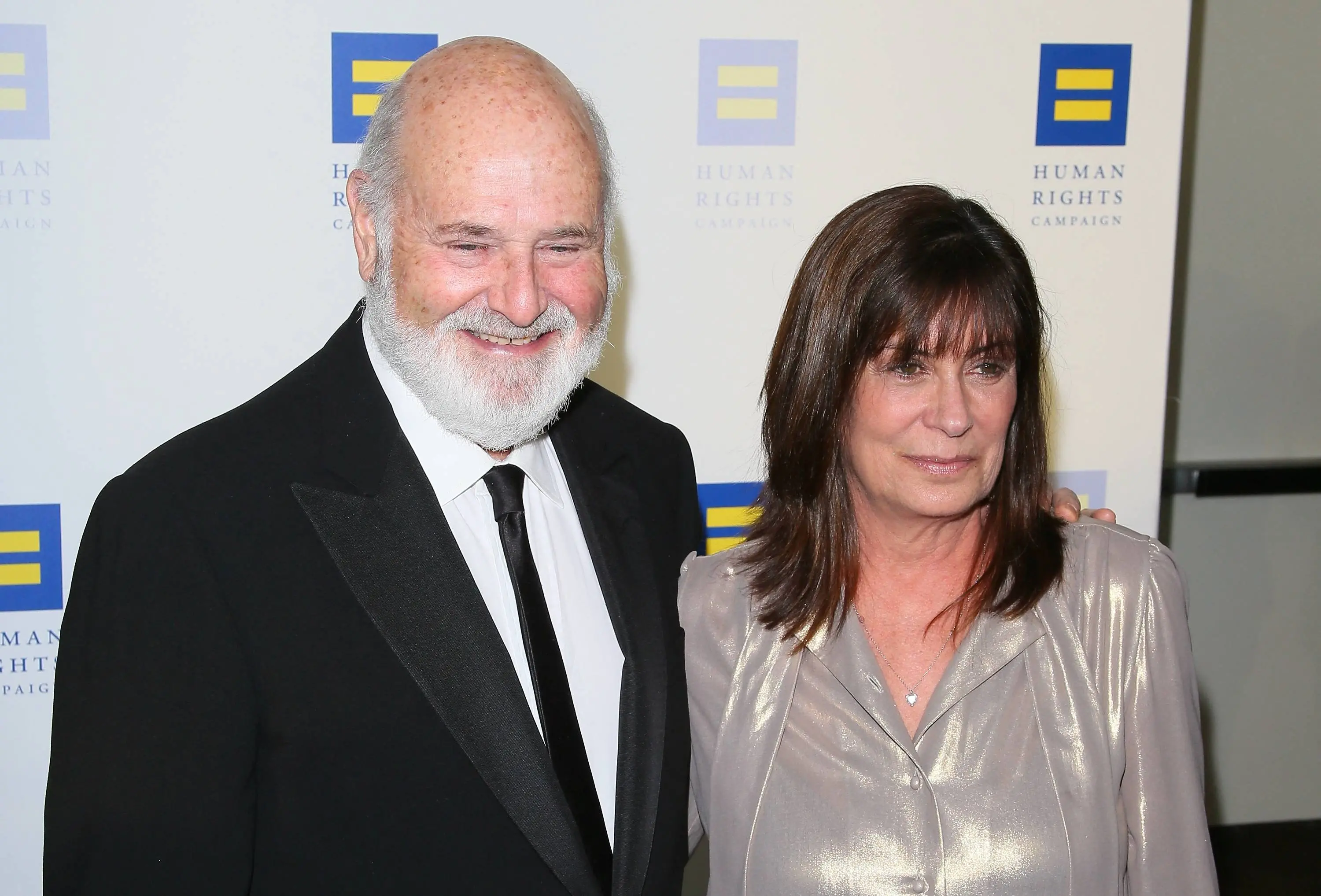 Imagem mostra o diretor Rob Reiner, com barba branca e smoking, e sua esposa Michele Singer Reiner, de blusa brilhante, posam para foto em gala.