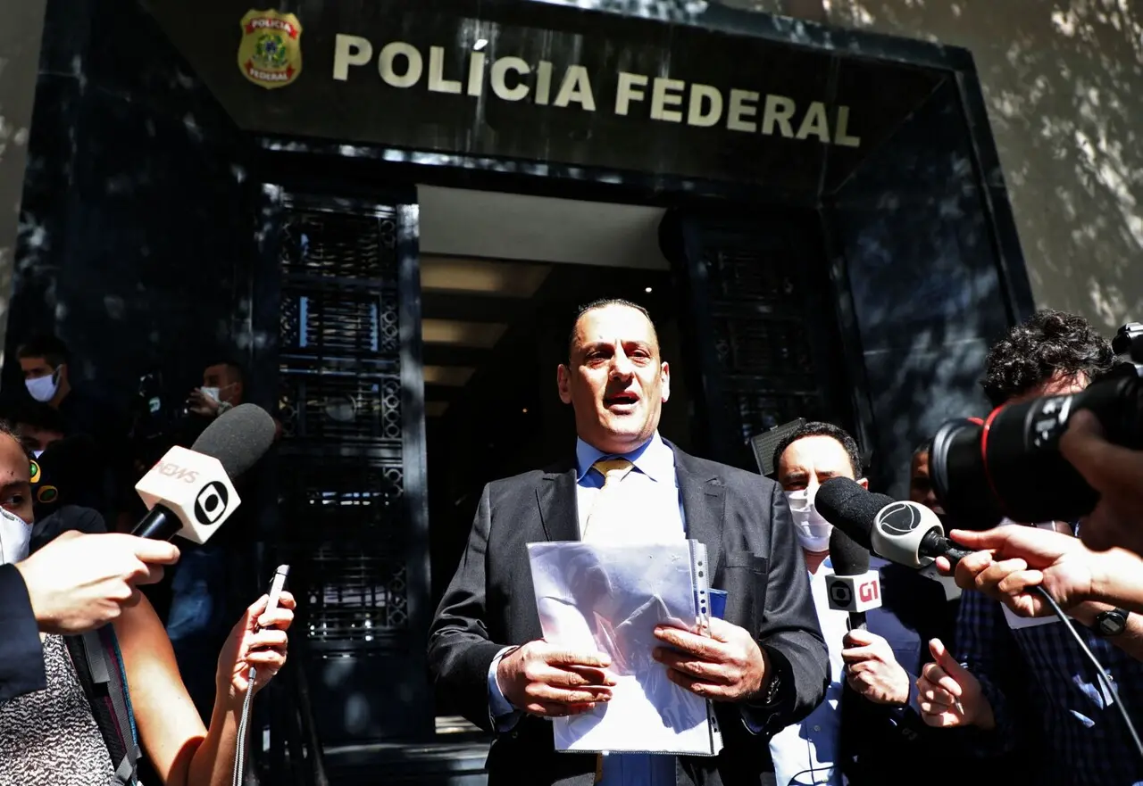 Homem branco de cabelos pretos vestindo um terno preto e branco. Ele está em pé, com um papel em mãos, cercado por microfones. Atrás, há o símbolo e os dizeres da Polícia Federal, junto à entrada do estabelecimento, em arquitetura preta.
