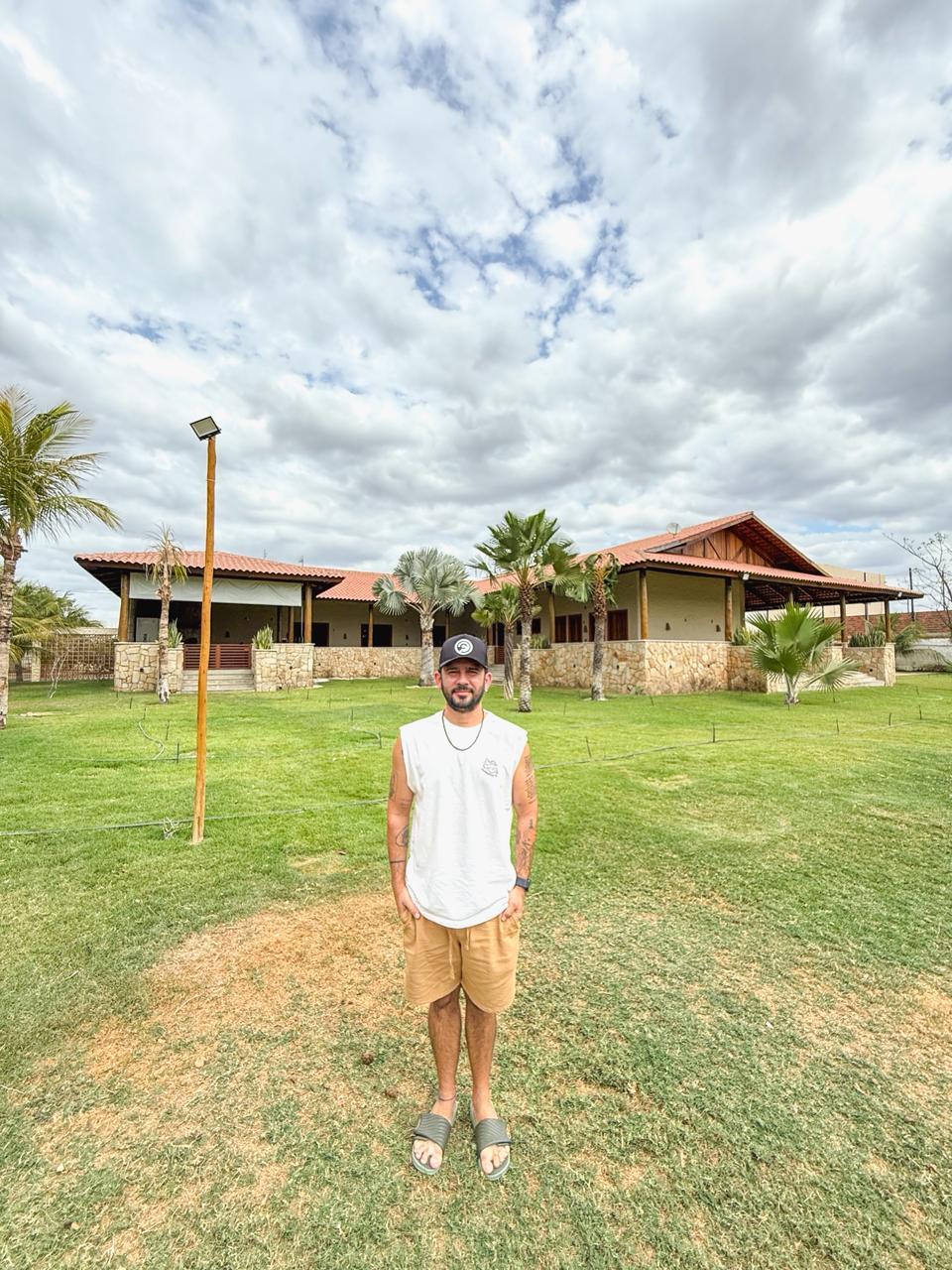 Na imagem, um homem jovem, com barba curta, veste camiseta regata branca, bermuda bege e boné preto, posando de pé no centro de um amplo gramado verde. Ele está à frente de uma casa de campo de um único pavimento, com arquitetura rústica, telhado de telhas de barro, colunas de madeira e base revestida em pedras naturais. O céu está nublado com nuvens claras e acinzentadas. Ao fundo e nas laterais da casa, veem-se palmeiras e coqueiros sob uma luz diurna suave.