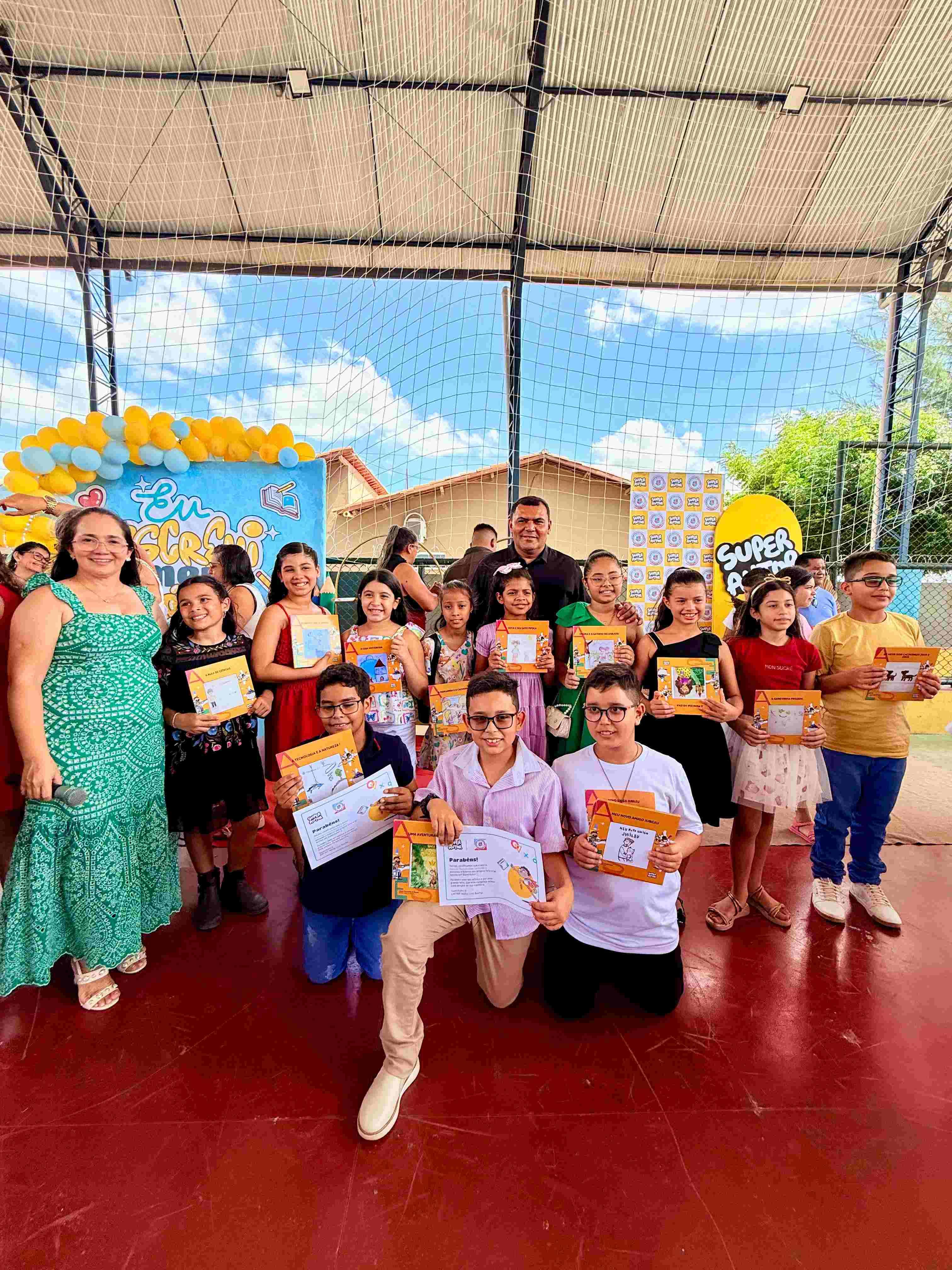 Imagem de estudantes do 4º e 5º ano da Escola Municipal Naiza Lira Rocha posam para foto durante o Super Evento de Autógrafos. As crianças aparecem segurando seus livros autorais e certificados de participação, acompanhadas por professores e integrantes da comunidade escolar. O espaço está decorado com painéis e balões que remetem ao projeto SuperAutor, marcando o lançamento coletivo das obras produzidas ao longo do ano letivo.