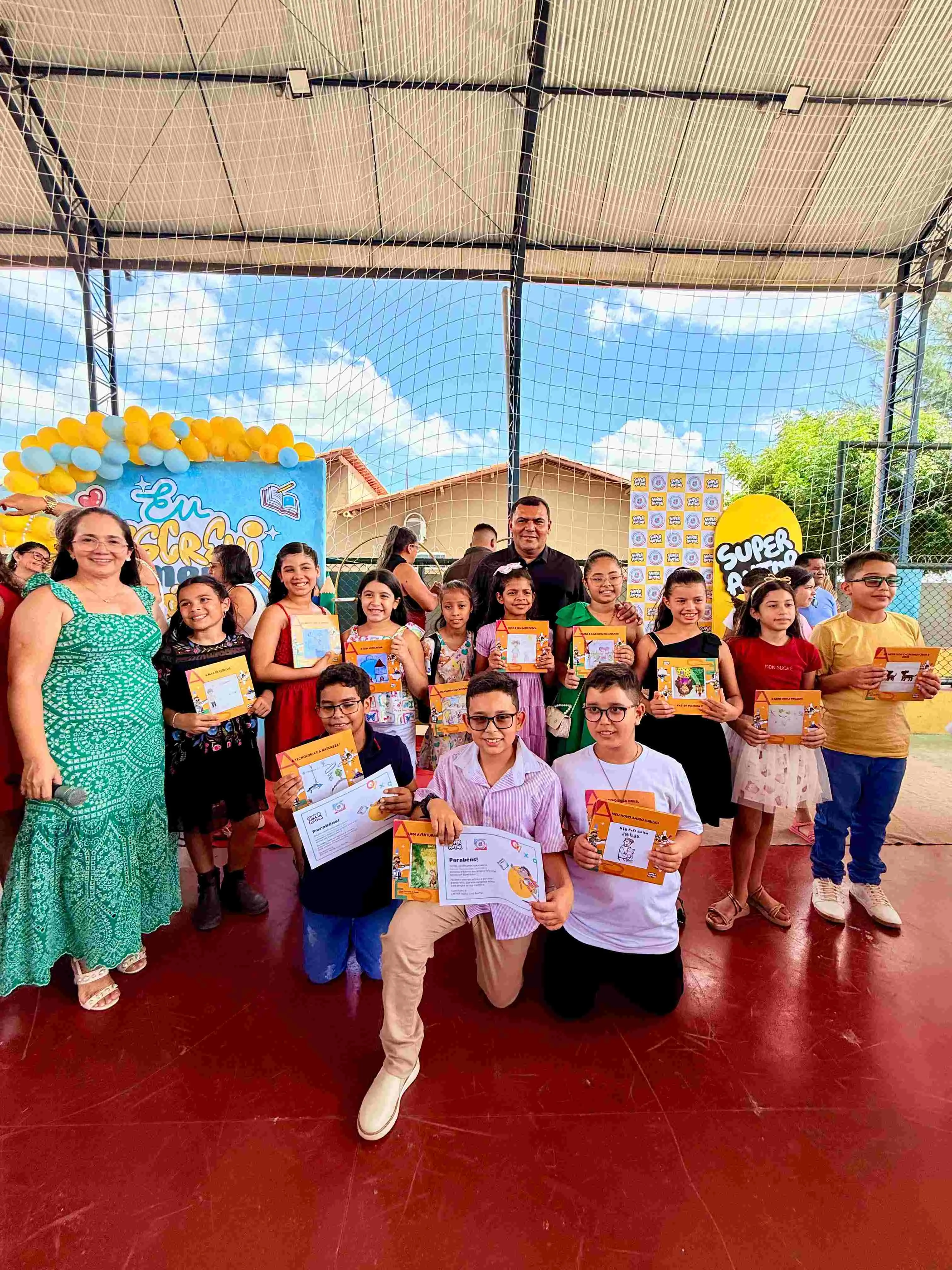 Imagem de estudantes do 4º e 5º ano da Escola Municipal Naiza Lira Rocha posam para foto durante o Super Evento de Autógrafos. As crianças aparecem segurando seus livros autorais e certificados de participação, acompanhadas por professores e integrantes da comunidade escolar. O espaço está decorado com painéis e balões que remetem ao projeto SuperAutor, marcando o lançamento coletivo das obras produzidas ao longo do ano letivo.