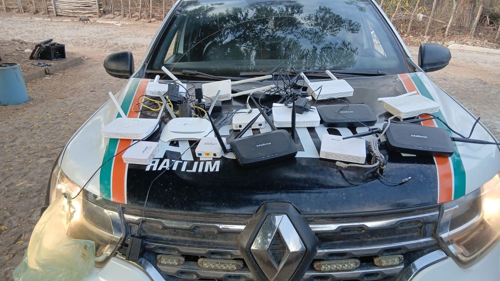 Diversos roteadores, modems e cabos eletrônicos estão dispostos sobre o capô de uma viatura da polícia, que está estacionada em uma área rural com chão de terra e vegetação ao fundo.