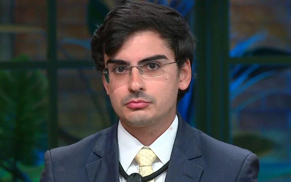 Pessoa vestindo terno escuro, camisa branca e gravata clara fala diante de um microfone, em um ambiente interno com iluminação suave e elementos decorativos ao fundo.