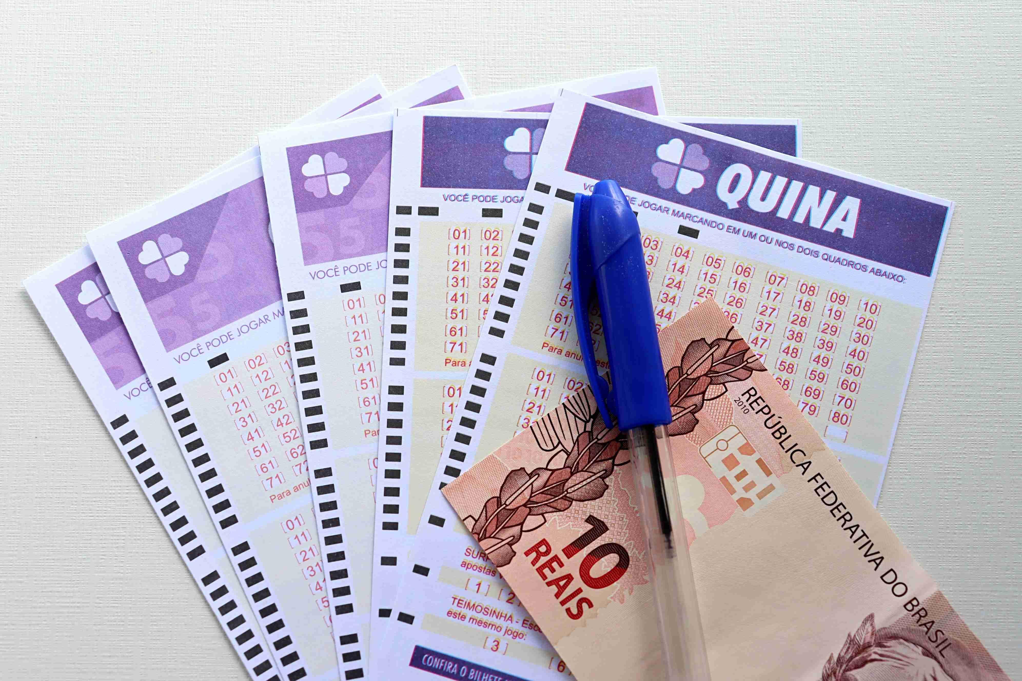 Imagem mostra bilhetes da loteria Quina, uma caneta azul e uma nota de dez reais sobre uma superfície branca.