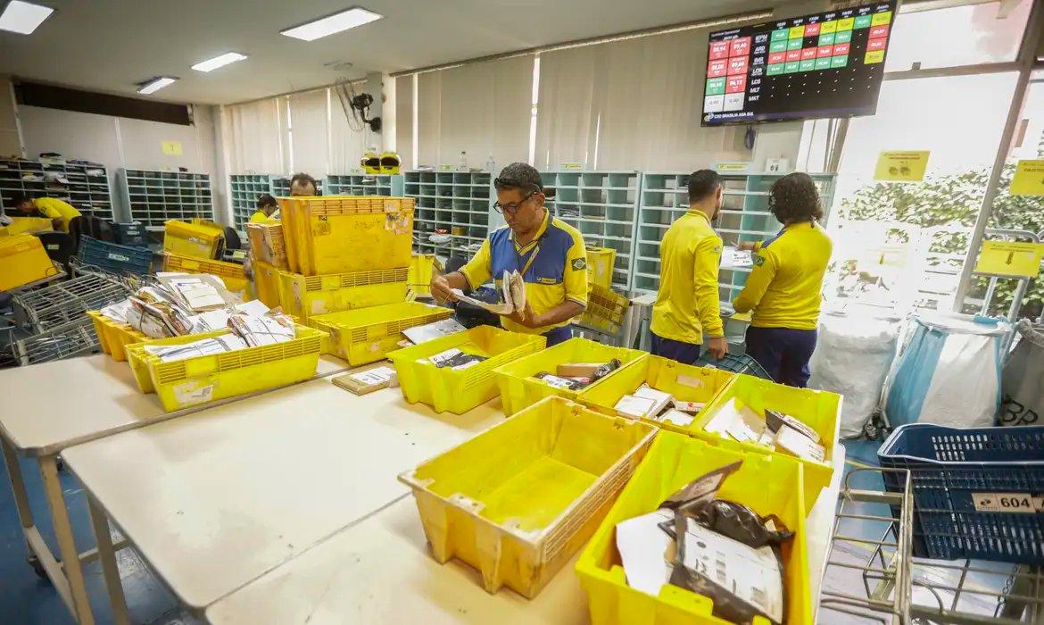 Funcionários dos Correios em uniformes amarelos e azuis, organizando cartas e pacotes em um centro de distribuição movimentado.