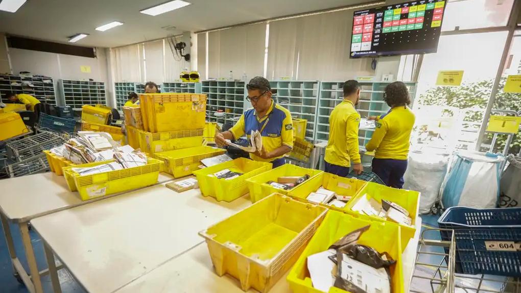 Funcionários dos Correios em uniformes amarelos e azuis, organizando cartas e pacotes em um centro de distribuição movimentado.
