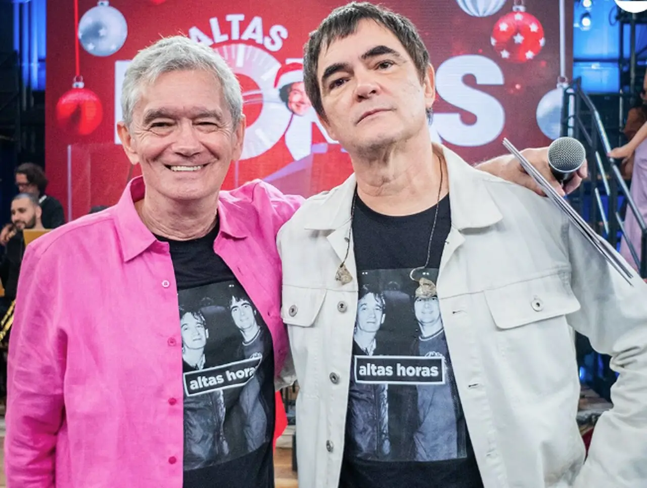 Dois homens brancos lado a lado sorrindo. À esquerda, ele tem cabelos brancos e usa uma blusa preta com a foto de ambos os homens mais jovens e a logo 