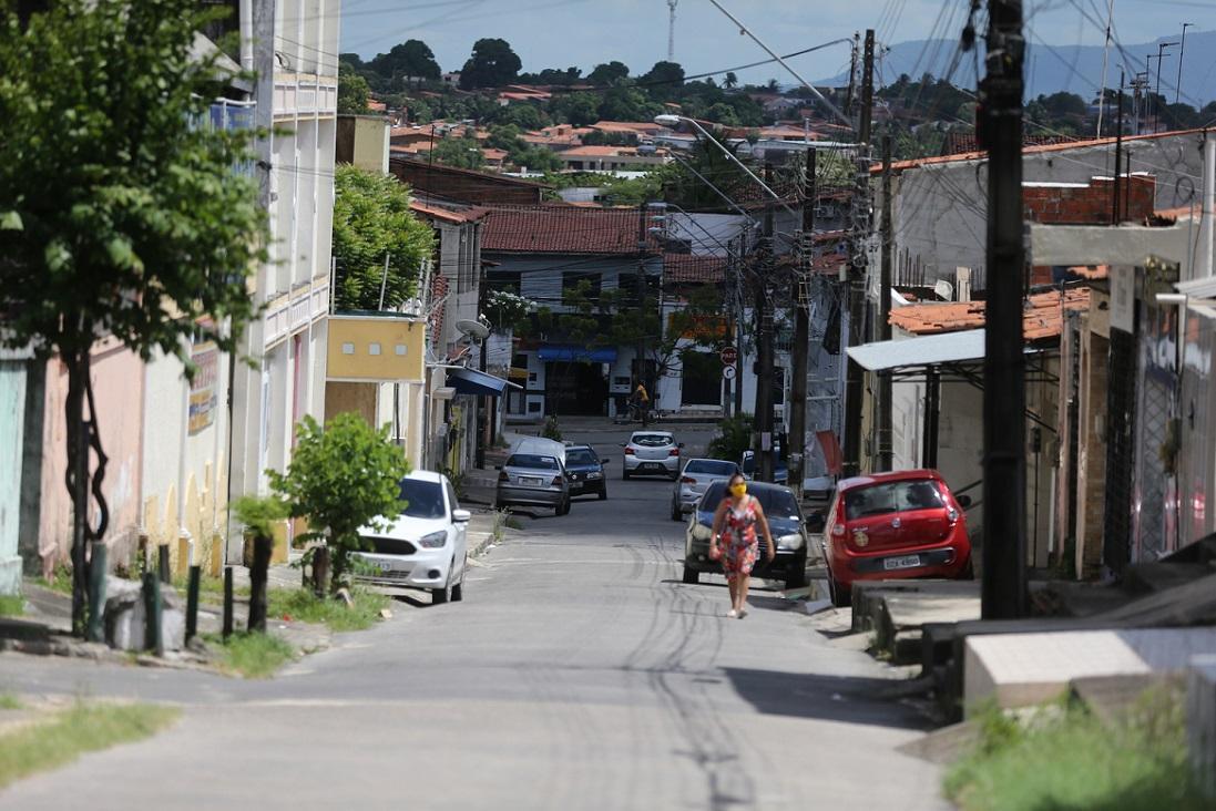 Imagem mostra rua de bairro em Fortaleza, com casas e carros estacionados. Uma mulher caminha na rua.
