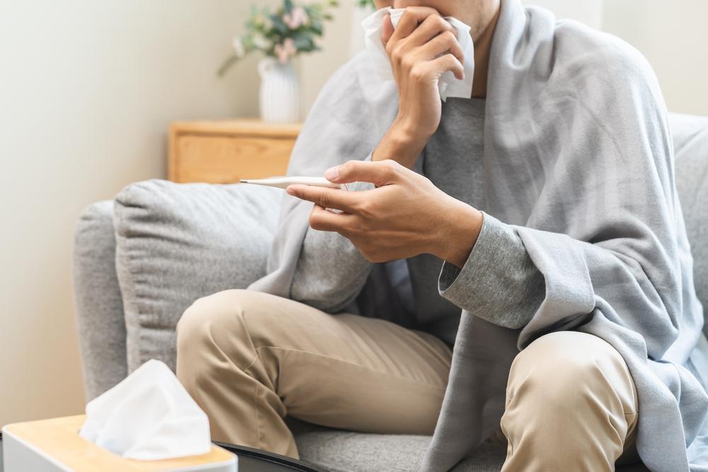 Pessoa sentada no sofá, coberta com manta, segurando termômetro e lenço, com caixa de lenços à frente.
