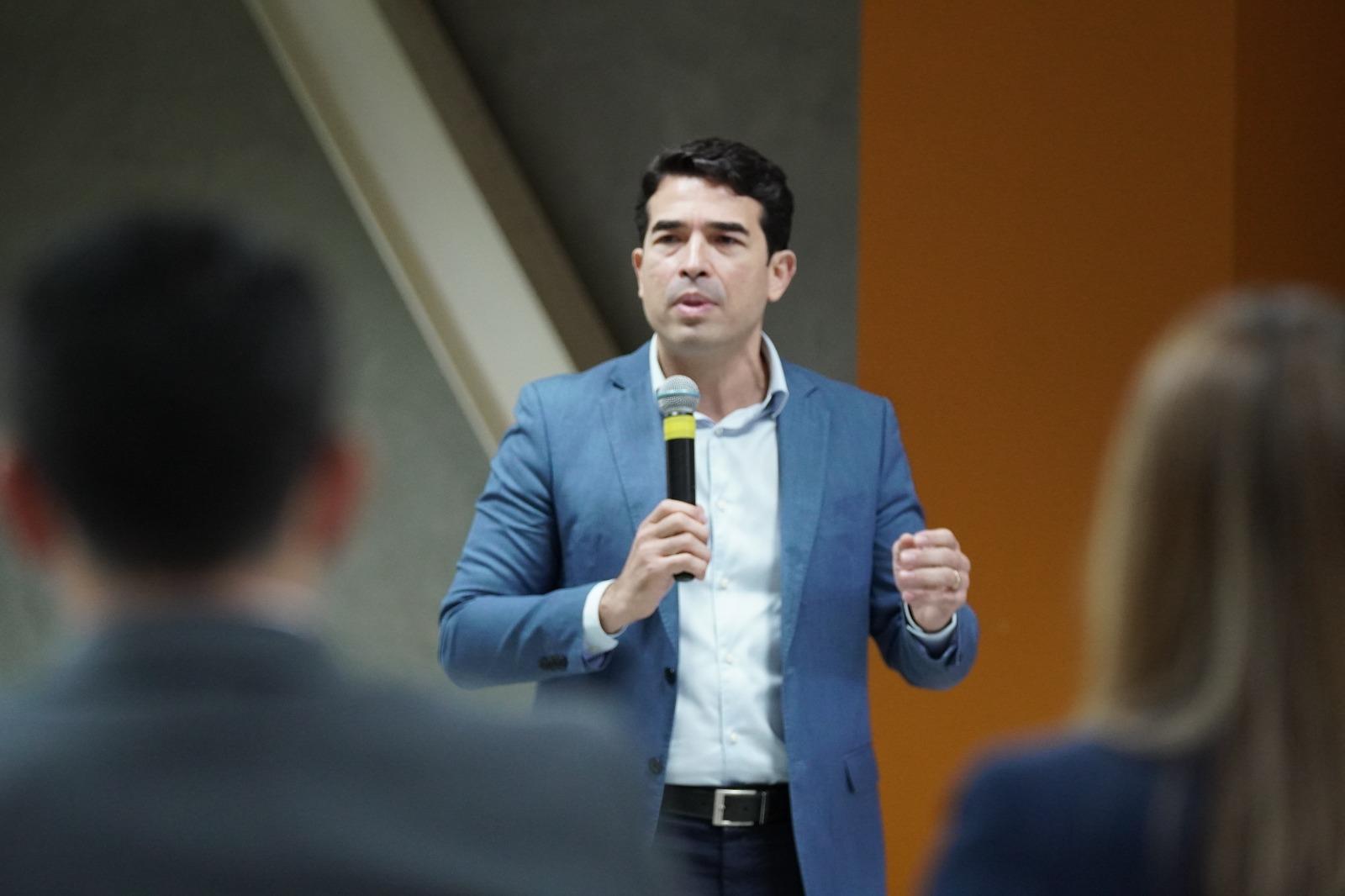 Rholden Queiroz, presidente do TCE Ceará, de terno azul e camisa branca segura um microfone e fala em pé para uma plateia, em ambiente interno, com pessoas desfocadas em primeiro plano.