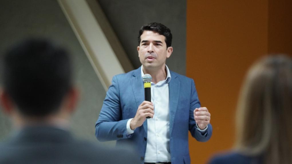 Rholden Queiroz, presidente do TCE Ceará, de terno azul e camisa branca segura um microfone e fala em pé para uma plateia, em ambiente interno, com pessoas desfocadas em primeiro plano.