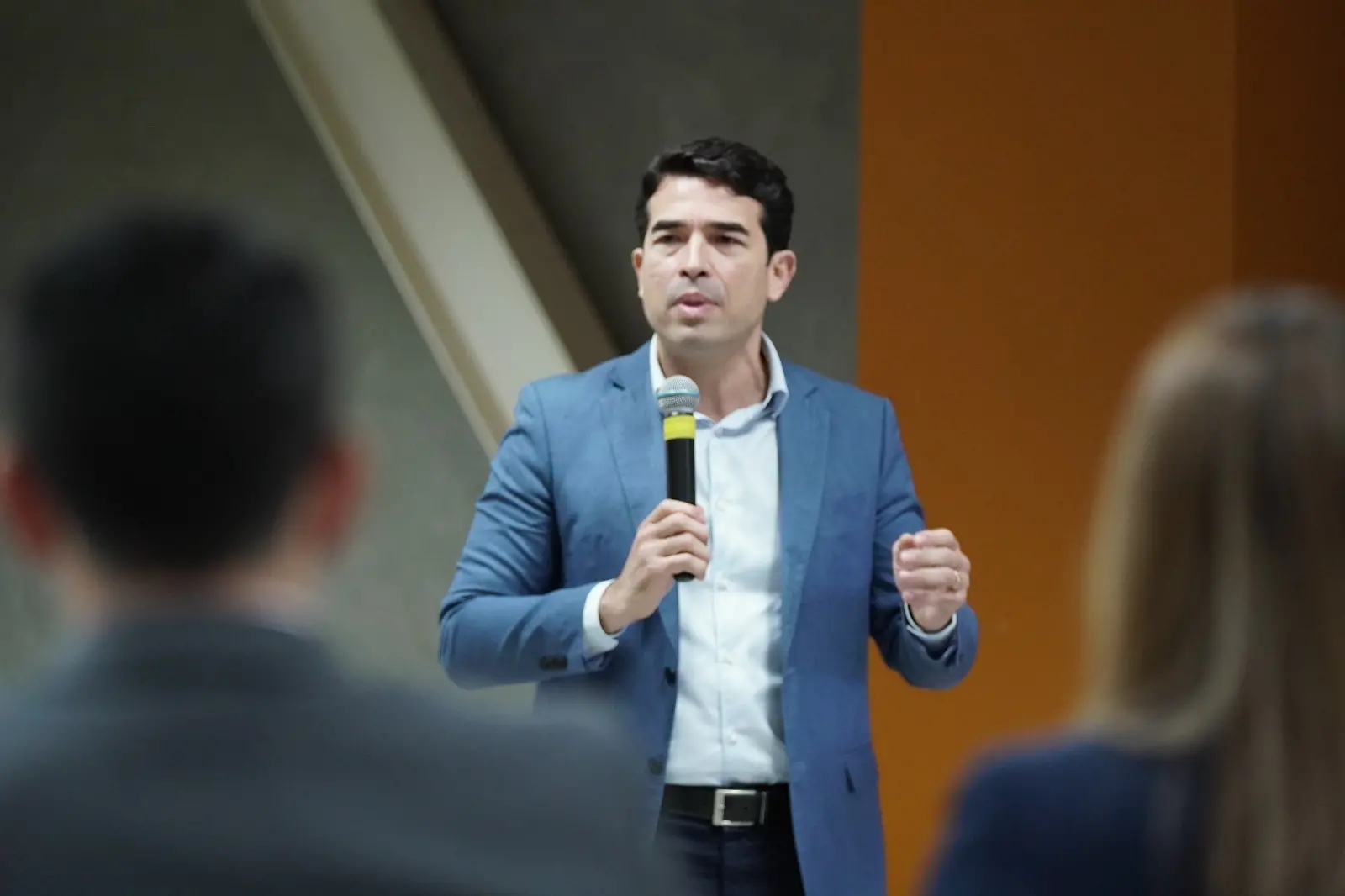 Rholden Queiroz, presidente do TCE Ceará, de terno azul e camisa branca segura um microfone e fala em pé para uma plateia, em ambiente interno, com pessoas desfocadas em primeiro plano.