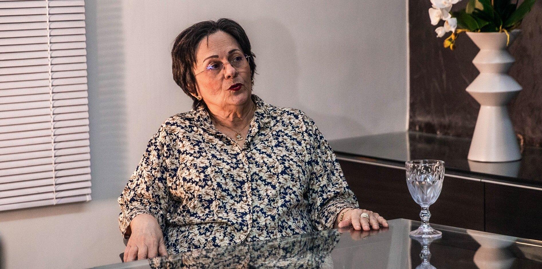 Uma pessoa sentada à mesa de vidro em um ambiente interno, usando uma blusa estampada de mangas compridas. Suas mãos repousam sobre a mesa. Ao lado direito, há uma taça decorativa transparente e um vaso branco alto com flores brancas. Ao fundo, aparece uma parede com persianas e um painel escuro, compondo um ambiente moderno e bem iluminado.