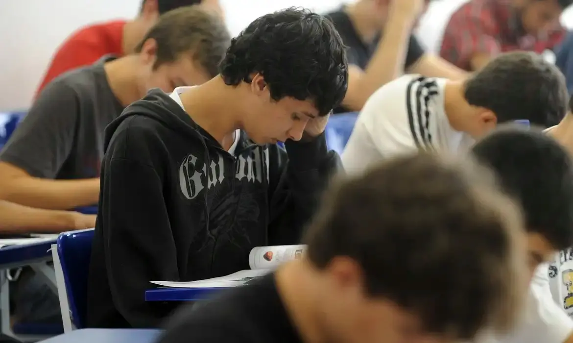 Alunos em ambiente escolar, com um candidato a concurso em primeiro plano lendo ou escrevendo em sua mesa.